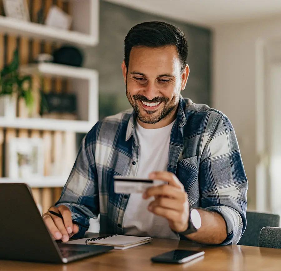 Um homem moreno jovem, sorri para um cartão de crédito. À sua frente encontra-se um caderno de apontamentos e um computador portátil. Um homem moreno jovem, sorri para um cartão de crédito. À sua frente encontra-se um caderno de apontamentos e um computador portátil.