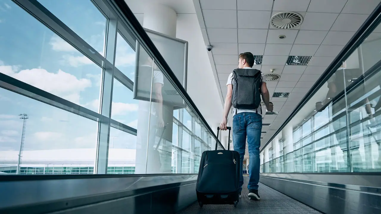 Homem a caminhar num tapete rolante dentro de um aeroporto, com uma mochila às costas e a puxar uma mala de viagem com rodas. O corredor tem janelas grandes com vista para o exterior, onde se vê o céu azul com algumas nuvens. Homem a caminhar num tapete rolante dentro de um aeroporto, com uma mochila às costas e a puxar uma mala de viagem com rodas. O corredor tem janelas grandes com vista para o exterior, onde se vê o céu azul com algumas nuvens.