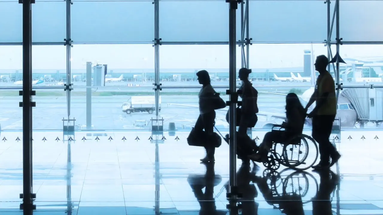 Imagem em contraluz de quatro pessoas a deslocarem-se num aeroporto. Duas delas encontram-se a carregar a sua bagagem, outra encontra-se numa cadeira de rodas que está a ser guiada pela última pessoa. Imagem em contraluz de quatro pessoas a deslocarem-se num aeroporto. Duas delas encontram-se a carregar a sua bagagem, outra encontra-se numa cadeira de rodas que está a ser guiada pela última pessoa.