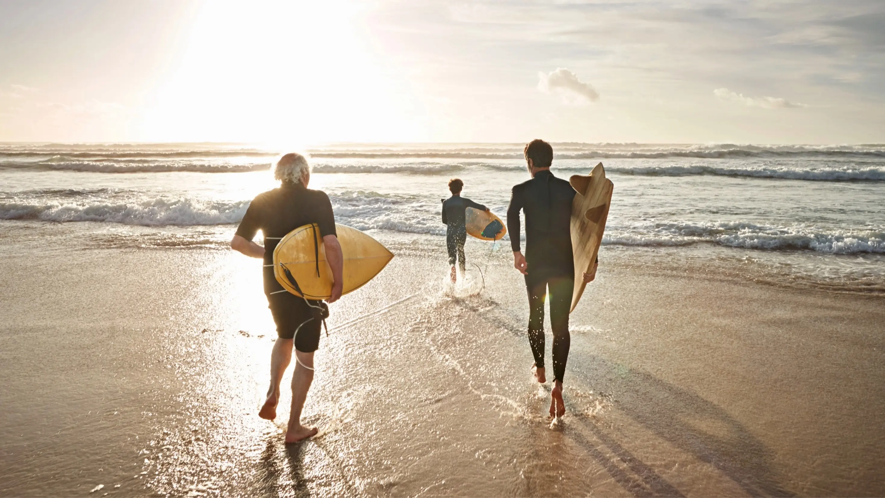 Três pessoas, cada uma delas com a sua prancha e fato de surf, prontaspara entrar no mar numa praia. Do lado esquerdo pode observar-se o reflexo do sol no mar e na areia. Três pessoas, cada uma delas com a sua prancha e fato de surf, prontaspara entrar no mar numa praia. Do lado esquerdo pode observar-se o reflexo do sol no mar e na areia.