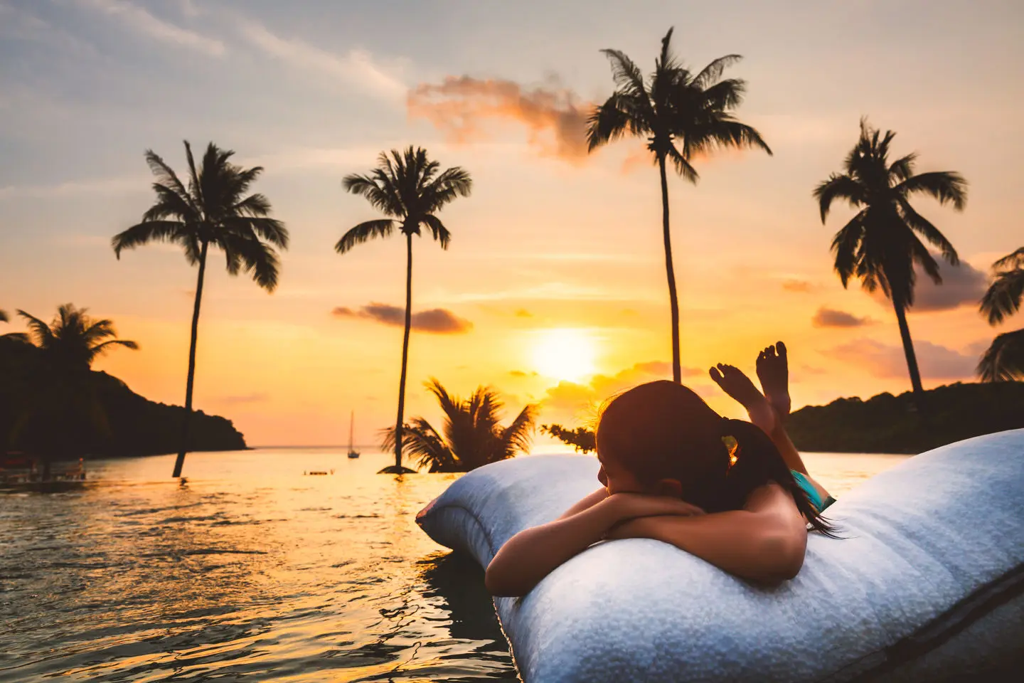 Uma senhora encontra-se deitada sobre uma almofada insuflável a flutuar na piscina de hotel de um local paradisíaco. No plano de fundo da imagem, observa-se o mar, várias palmeiras e o por-do sol. Uma senhora encontra-se deitada sobre uma almofada insuflável a flutuar na piscina de hotel de um local paradisíaco. No plano de fundo da imagem, observa-se o mar, várias palmeiras e o por-do sol.